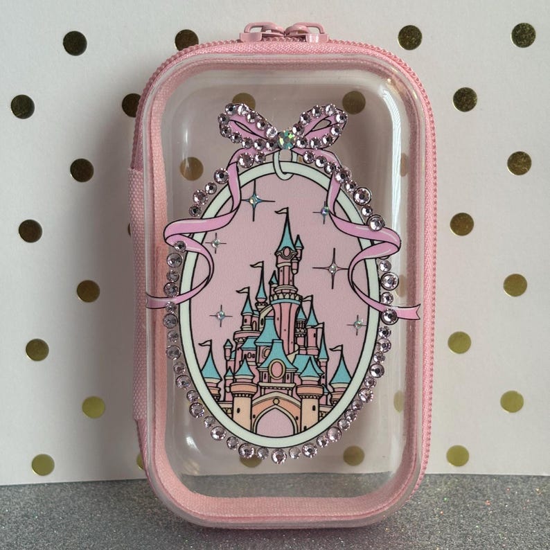 May include: A pink, clear, zippered case with a castle design. The case features a pink bow and an oval frame with a castle illustration. The frame and bow are embellished with rhinestones. The case is designed to hold small items.