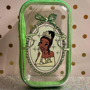 May include: Clear, rectangular cosmetic bag with a green zip and trim. The bag features a framed illustration of a princess in a green dress and tiara, surrounded by green rhinestones and a bow.