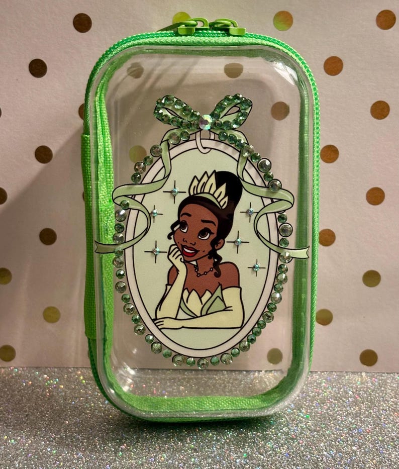May include: Clear, rectangular cosmetic bag with a green zip and trim. The bag features a framed illustration of a princess in a green dress and tiara, surrounded by green rhinestones and a bow.