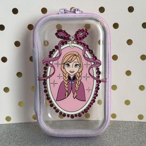 May include: A clear, rectangular zipped case with a lilac trim. The case features a portrait of a cartoon character with blonde braids, wearing a purple cape, and surrounded by a pink oval frame with rhinestones and a bow.