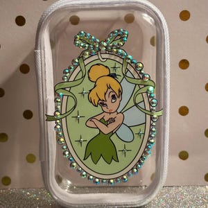 May include: A clear, zipped pouch with white trim. It features a cartoon image of Tinkerbell in a green dress, within a decorative oval frame with rhinestones and a green ribbon. The background is a light green colour.