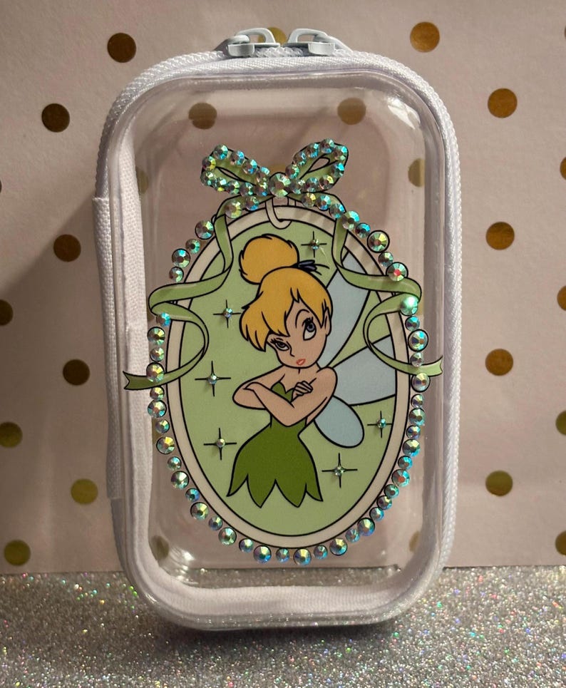 May include: A clear, zipped pouch with white trim. It features a cartoon image of Tinkerbell in a green dress, within a decorative oval frame with rhinestones and a green ribbon. The background is a light green colour.