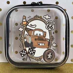 May include: Clear, square zippered case featuring a cartoon image of Mater from the movie Cars, framed by a decorative border. The design includes a Route 66 sign, clouds, and checkered flags. The case has a black trim.