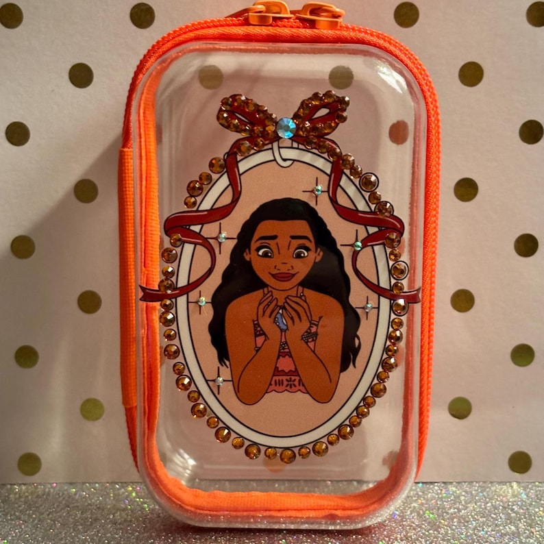 May include: A clear, rectangular cosmetic bag with an orange zip and trim. The bag features a cartoon image of a woman in an oval frame decorated with rhinestones and a bow. The bag is designed to hold makeup or other small items.