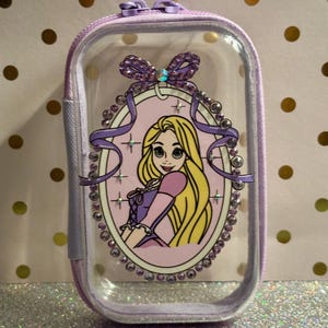 May include: A clear, rectangular cosmetic bag with a purple zip and trim. The bag features a framed image of a cartoon princess with long blonde hair, wearing a pink and purple dress. The frame is decorated with rhinestones and a purple bow.