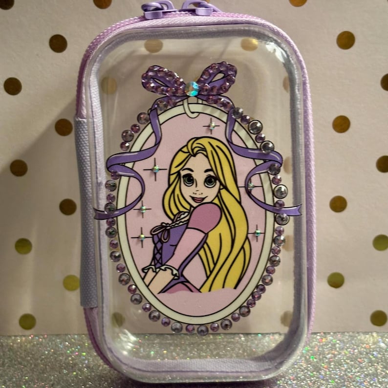 May include: A clear, rectangular cosmetic bag with a purple zip and trim. The bag features a framed image of a cartoon princess with long blonde hair, wearing a pink and purple dress. The frame is decorated with rhinestones and a purple bow.