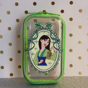 May include: Clear rectangular case with a green zip and trim. The case features a cartoon image of a woman in a green and blue outfit, holding a lantern. The image is surrounded by a decorative oval with green gems and a bow.