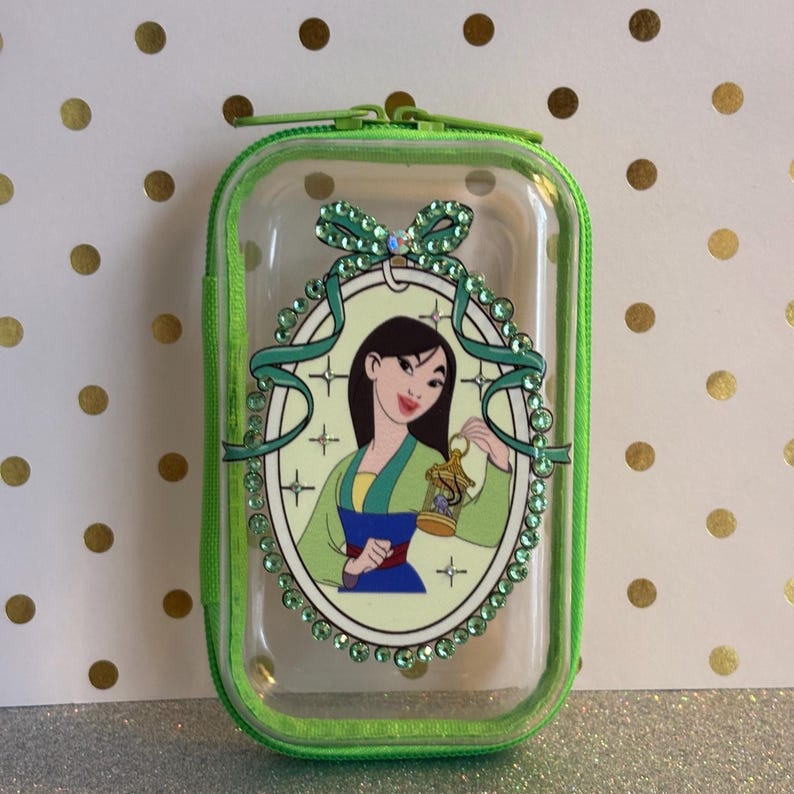 May include: Clear rectangular case with a green zip and trim. The case features a cartoon image of a woman in a green and blue outfit, holding a lantern. The image is surrounded by a decorative oval with green gems and a bow.
