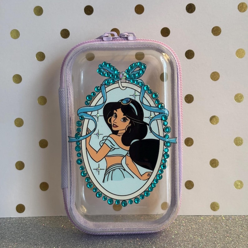 May include: A small, lavender-coloured zippered case with a clear front panel. The panel features a cartoon image of a princess in a blue outfit, framed by a blue ribbon and sparkling embellishments. The case has a small handle.