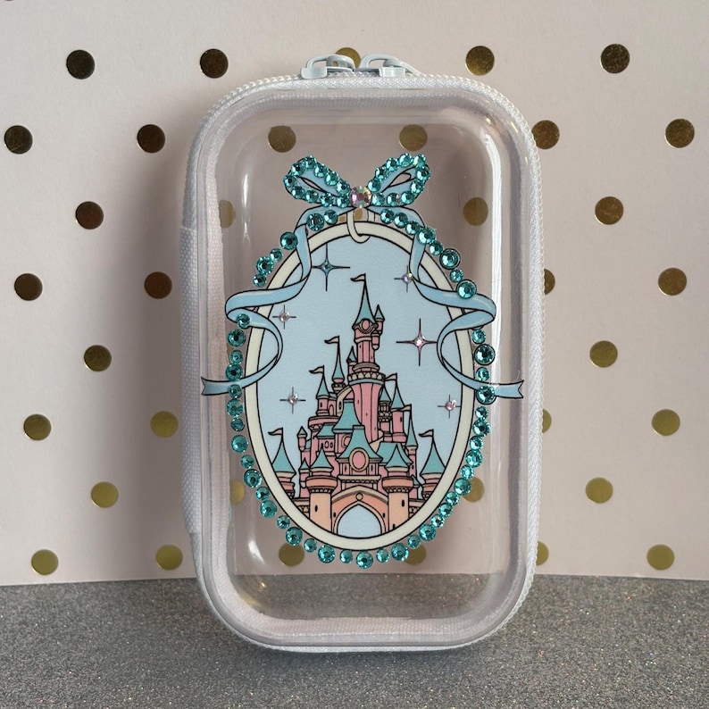 May include: Clear zipped case with a castle design in pink, blue, and beige. The castle is framed by a light blue oval with a bow and sparkling embellishments. The case has a white zip and is on a gold polka dot background.