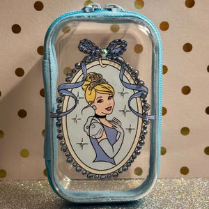 May include: A clear, rectangular cosmetic bag with a light blue zip and trim. The bag features a Cinderella illustration within a light blue oval frame, adorned with rhinestones and a bow. The background is a glittery surface.