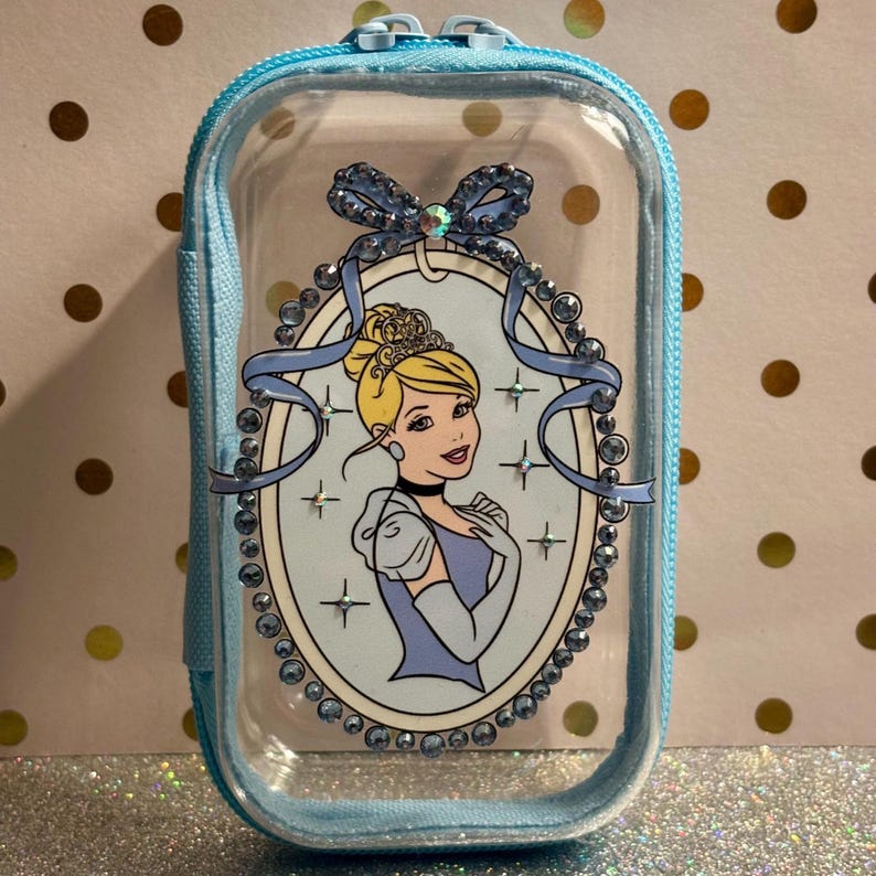 May include: A clear, rectangular cosmetic bag with a light blue zip and trim. The bag features a Cinderella illustration within a light blue oval frame, adorned with rhinestones and a bow. The background is a glittery surface.