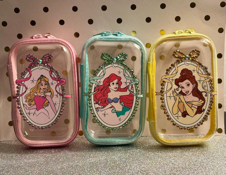 May include: Three clear, rectangular zipped pouches with princess illustrations. The pouches are pink, teal, and yellow, each featuring a different princess in an oval frame with decorative embellishments. The pouches have a clear front and a solid colour border.