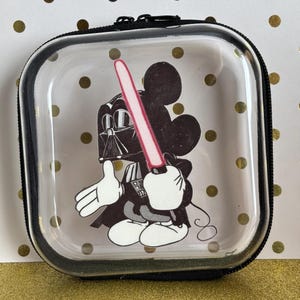May include: A clear, square case with a black zipper and trim. The case features a cartoon illustration of Mickey Mouse dressed as Darth Vader, holding a pink lightsaber. The background has gold polka dots.