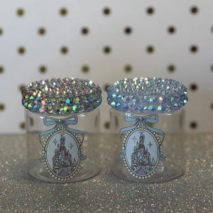 May include: Two clear cylindrical jars with lids covered in iridescent rhinestones. Each jar features a light blue oval decal depicting a castle illustration framed by a decorative border and topped with a bow graphic, creating a cohesive set.