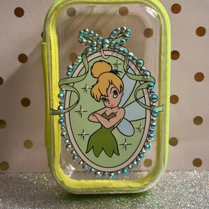May include: A clear, rectangular cosmetic bag with a neon yellow zip and trim. The bag features a cartoon image of Tinkerbell in a green oval frame, embellished with iridescent rhinestones. The background is a light green colour.