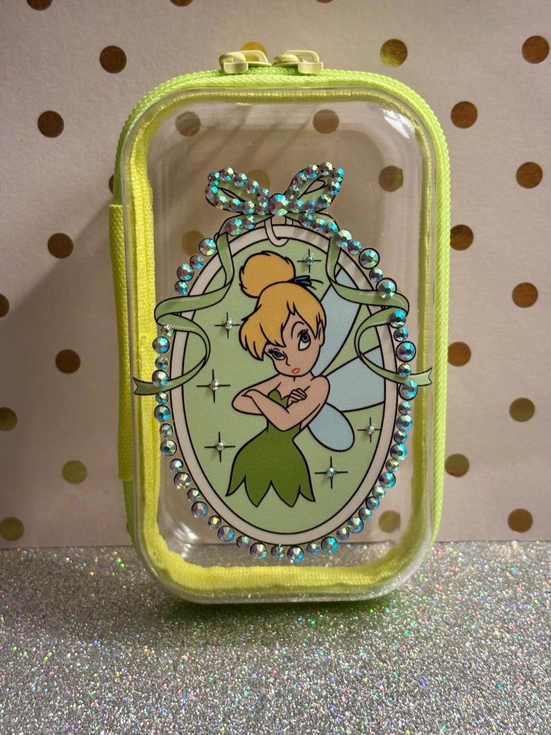 May include: A clear, rectangular cosmetic bag with a neon yellow zip and trim. The bag features a cartoon image of Tinkerbell in a green oval frame, embellished with iridescent rhinestones. The background is a light green colour.