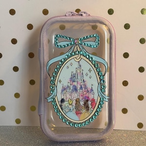 May include: A clear, rectangular case with a lavender zip and trim. The case features a decorative oval with a castle illustration, surrounded by teal rhinestones and a teal bow. The case is designed for small items.