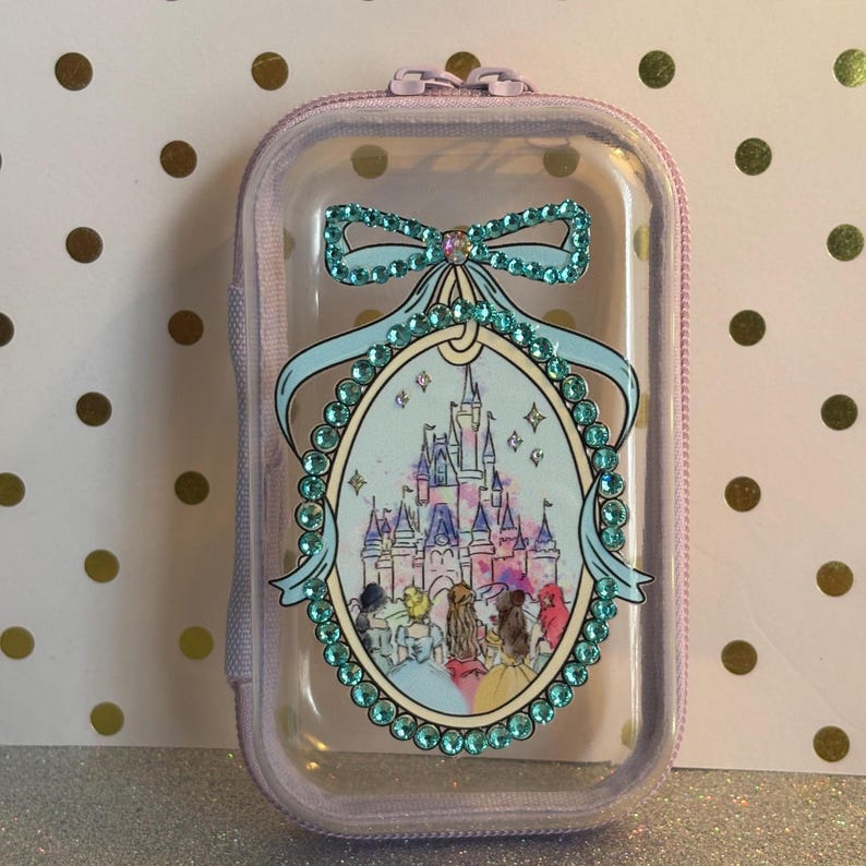 May include: A clear, rectangular case with a lavender zip and trim. The case features a decorative oval with a castle illustration, surrounded by teal rhinestones and a teal bow. The case is designed for small items.