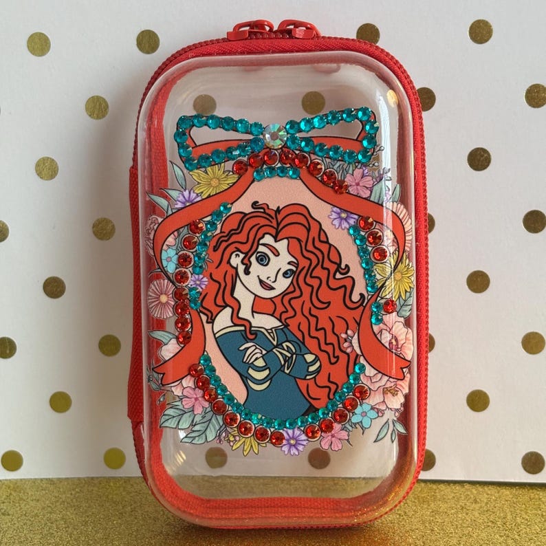 May include: A red zipped case with a clear front panel. The panel features a cartoon illustration of a character with red hair, framed by flowers and jewels. The case has a red zip and a red carrying strap.