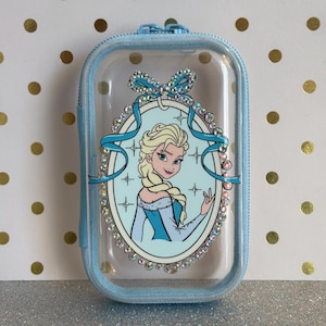 May include: A light blue, rectangular zipped case with a clear front. The case features a cartoon image of Elsa from Frozen, surrounded by a decorative oval frame with rhinestones and a blue bow.