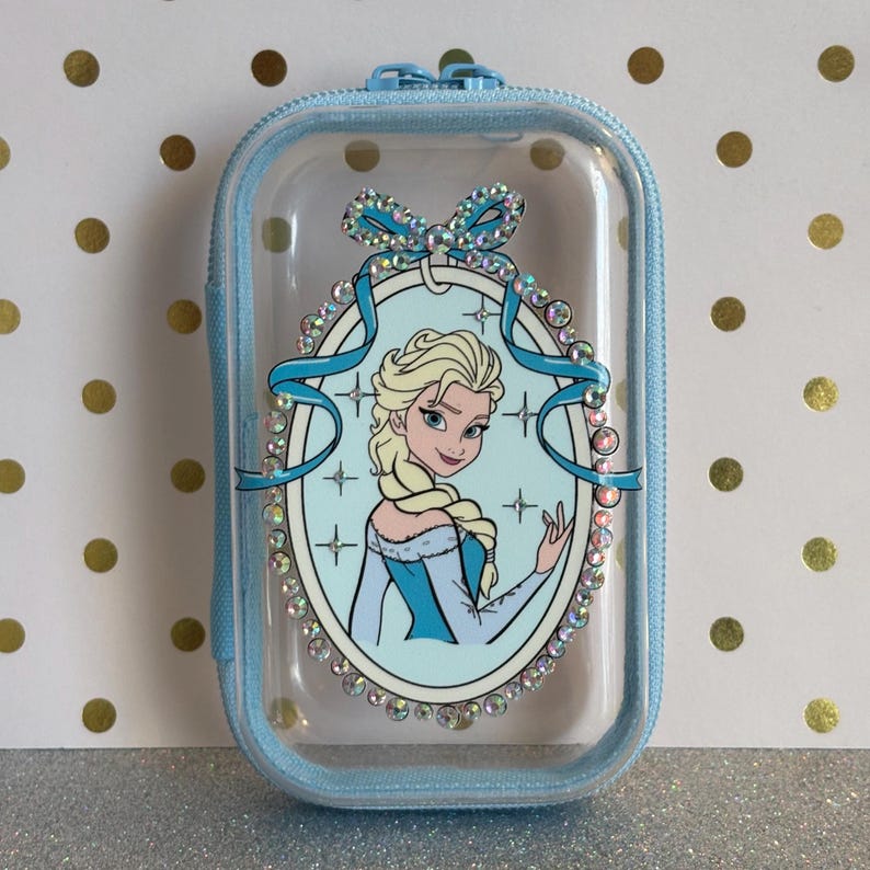 May include: A light blue, rectangular zipped case with a clear front. The case features a cartoon image of Elsa from Frozen, surrounded by a decorative oval frame with rhinestones and a blue bow.