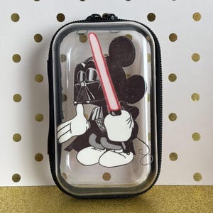 May include: A clear, rectangular case with a black zipper and a black border. Inside, a cartoon image of Mickey Mouse dressed as Darth Vader, holding a red lightsaber. The background is a gold and white polka dot pattern.