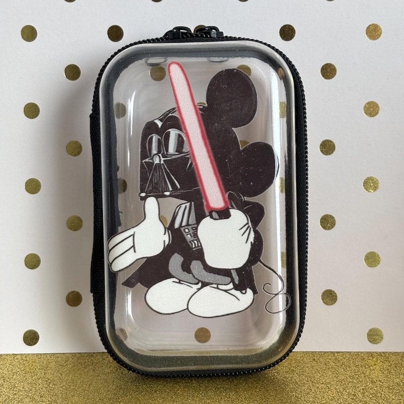May include: A clear, rectangular case with a black zipper and a black border. Inside, a cartoon image of Mickey Mouse dressed as Darth Vader, holding a red lightsaber. The background is a gold and white polka dot pattern.