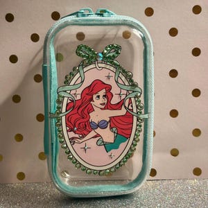 May include: A clear, rectangular cosmetic bag with a teal zip and trim. The bag features a cartoon image of Ariel from The Little Mermaid, surrounded by a decorative oval frame with rhinestones and a bow.