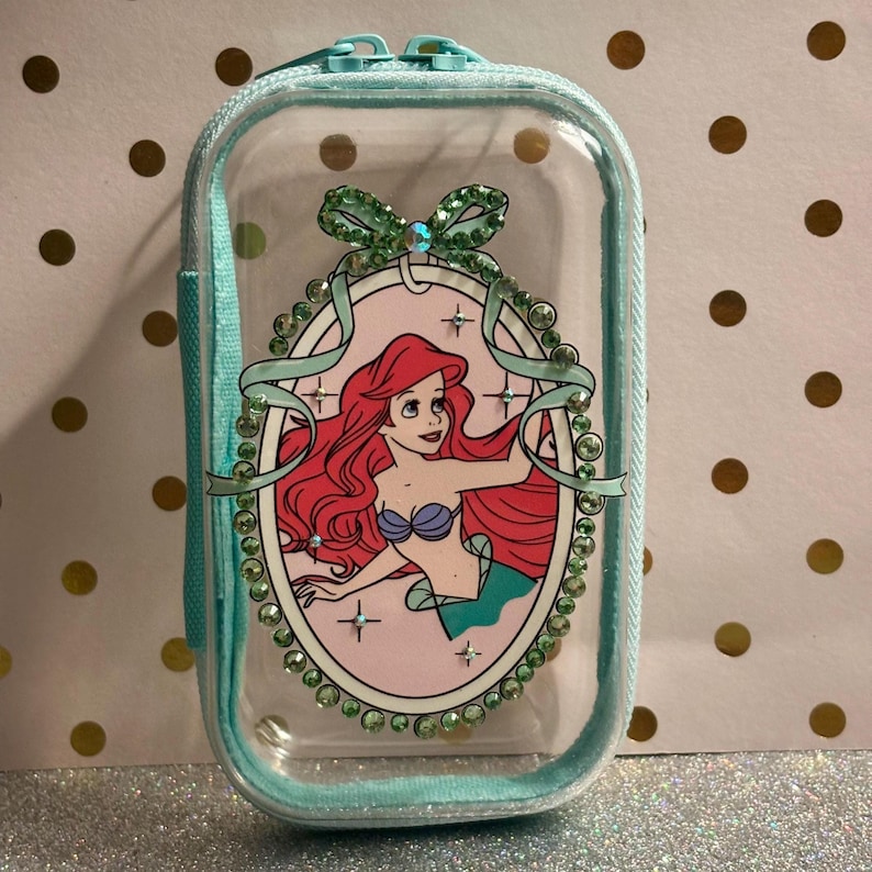 May include: A clear, rectangular cosmetic bag with a teal zip and trim. The bag features a cartoon image of Ariel from The Little Mermaid, surrounded by a decorative oval frame with rhinestones and a bow.