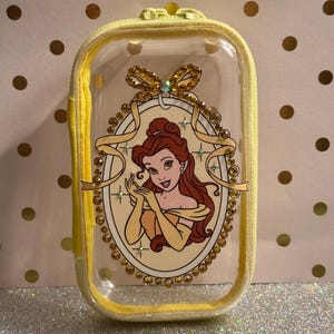 May include: A clear, rectangular cosmetic bag with a yellow zip and trim. The bag features a cartoon image of Belle from Beauty and the Beast, surrounded by gold-coloured embellishments. The bag is approximately 13 cm long.