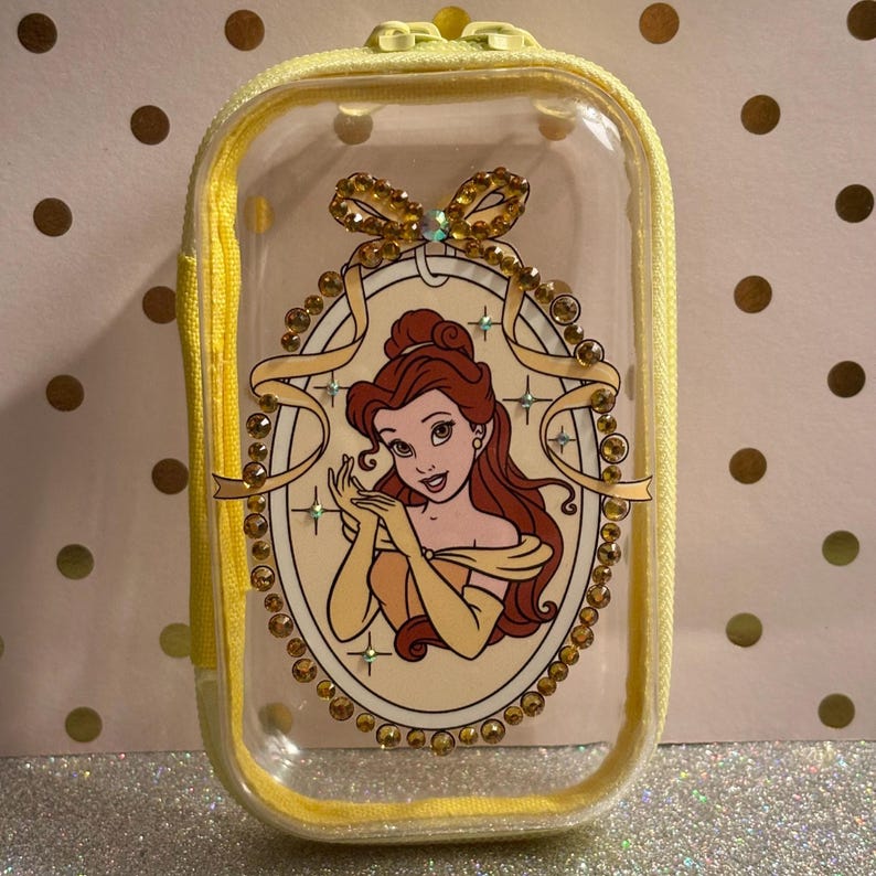 May include: A clear, rectangular cosmetic bag with a yellow zip and trim. The bag features a cartoon image of Belle from Beauty and the Beast, surrounded by gold-coloured embellishments. The bag is approximately 13 cm long.