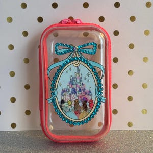 May include: A clear, rectangular case with a pink zip and trim. The case features a teal bow and a decorative oval with a castle and figures. The oval is outlined with teal gems.