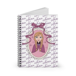 May include: A spiral-bound notebook featuring a cartoon illustration of a character with blonde braids in a pink oval frame. The frame is adorned with a purple ribbon and surrounded by the name "Anna" in a repeating pattern on a white background.
