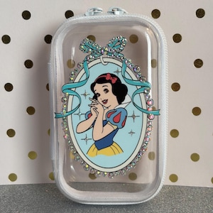 May include: A clear, zipped case with a Snow White graphic. The oval graphic is framed by rhinestones and a teal ribbon. The Snow White illustration has a red bow, blue and yellow dress, and black hair.