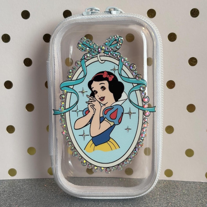 May include: A clear, zipped case with a Snow White graphic. The oval graphic is framed by rhinestones and a teal ribbon. The Snow White illustration has a red bow, blue and yellow dress, and black hair.