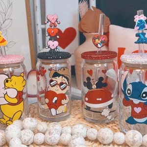 May include: Four clear glass tumblers with character designs, each with a lid and straw. The characters include Winnie the Pooh, a cartoon figure with heart-shaped sunglasses, Mickey and Minnie Mouse, and Stitch. The lids are decorated with glitter.