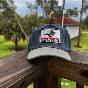 Op de afbeelding: Een baseballcap in denimstijl met een grijze klep en een rechthoekige patch. De patch toont het Winchester-logo met de tekst "Winchester Rifles and Shotguns". De pet ligt op een houten oppervlak.