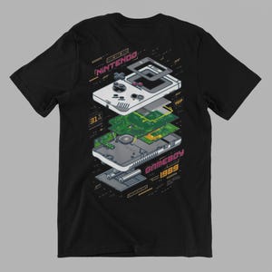 Può includere: T-shirt nera con un diagramma pixelato di un Nintendo Game Boy smontato. Il design include le parole "NINTENDO" e "GAMEBOY 1989" in un font retrò, con accenti verdi e rosa. La grafica mostra i componenti interni.