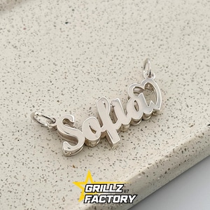 Puede incluir: Un colgante plateado con el nombre "Sofia" escrito en cursiva. El colgante tiene bucles para una cadena. El fondo es una superficie texturizada de color gris claro. El logotipo "GRILLZ FACTORY" está en la esquina inferior.