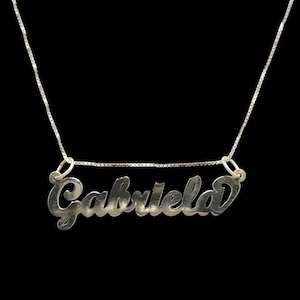 Puede incluir: Collar de plata con un colgante de placa de nombre cursivo que dice "Gabriela".
