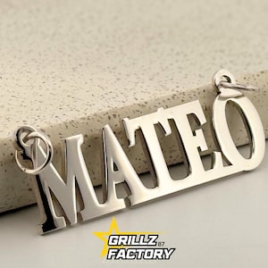 以下が含まれることがあります： 太字のサンセリフ体で「MATEO」という名前が刻まれたシルバーカラーのペンダント。ペンダントの両端には取り付け用のループが付いています。「GRILLZ 87 FACTORY」のロゴが下部にあります。