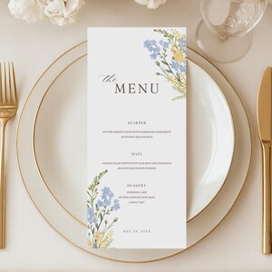Może przedstawiać: Pionowa biała karta menu z napisem "the MENU" oraz kwiatowymi ilustracjami niebieskich i żółtych polnych kwiatów. Menu zawiera listę przystawek, dań głównych i deserów, umieszczone na białym talerzu ze złotym brzegiem, w otoczeniu złotych sztućców.