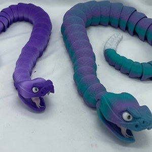 Puede incluir: Dos juguetes de serpiente segmentados de color morado y azul. Las serpientes están hechas de un material suave y flexible y tienen un diseño de serpiente realista. Las serpientes están enroscadas en una pose relajada.