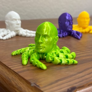 May include: Four 3D printed octopus figurines with human heads. The octopus figurines are in different colors: white, green, purple, and yellow. The octopus figurines are on a wooden surface.