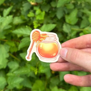 May include: A die-cut sticker featuring a clear glass jar filled with golden honey and a wooden honey dipper resting on the rim. The illustration uses warm yellow and orange tones with a white border, showcasing a glossy, smooth texture.