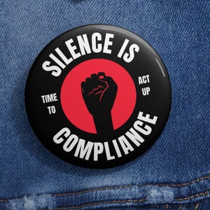 May include: A round black button with the text "SILENCE IS COMPLIANCE" in white, surrounding a red circle with a black fist. The button also includes the phrases "TIME TO" and "ACT UP". The button is pinned to a blue denim jacket.