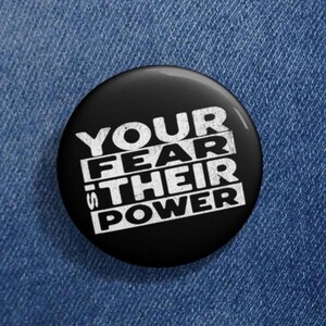 May include: Black button with white text that reads "Your fear is their power".