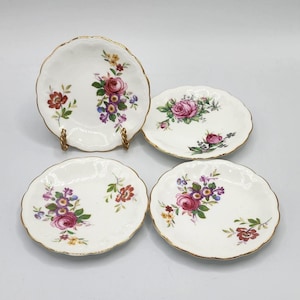 Może przedstawiać: Zestaw czterech białych porcelanowych spodków z pozłacanymi brzegami i kwiatowymi wzorami w odcieniach różowego, czerwonego i fioletowego.