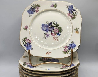 May include: A stack of ten square-shaped white porcelain plates with a gold rim and a floral design featuring blue, pink, and yellow flowers.